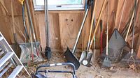 Full view of garden tools including shovels, rakes, post hole digger and axe resting against a wooden wall.
