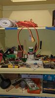 Photo of a shelf showing various tools, painting supplies, and electrical accessories such as extension cords and a surge protector. Small paint can and other small hardware items are visible.