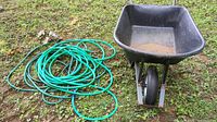 Photo showing black plastic wheelbarrow with metal frame and tire alongside two coiled green garden hoses on grass.
