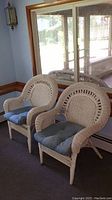 Two white wicker chairs with blue cushions positioned indoors near a window, reflecting natural daylight.