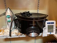 Front view of three items: Black Crockpot slow cooker, white and green Black and Decker iron, white Medline blood pressure monitor, all on shelf