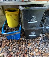 Photo showing two black recycling bins stacked and a yellow pail with mulch on a blue crate on gravel ground with leaves.