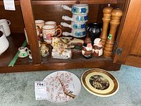 Display of ceramic and stoneware figurines, salt and pepper shakers, mugs, stacked bowls, wooden salt and pepper grinders, and metal holder on a wooden shelf.