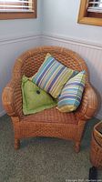 Full view of natural brown woven wicker armchair with three colorful decorative pillows (striped and solid green with button). Chair has curved back and arms, placed on carpet next to windows.