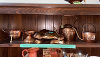 Photo of copper ware items displayed on wooden shelves including molds, teapot, candle holder, strainer, watering can and copper bowls.
