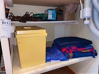Photo showing yellow plastic file box and blue and red woven blankets on a white shelf.