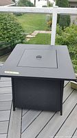 Square black metal propane fire pit with flat top and center handle, shown on outdoor deck.