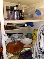 Two shelves of kitchen ware including blender parts, plastic storage containers, glass and ceramic serving ware, wooden bowls, Pyrex bakeware, flatware.