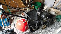 Craftsman lawn mower with Honda engine and attached black grass bagger, alongside red jerry can on gravel floor.