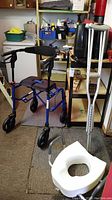 Photo showing the Drive white raised toilet seat on the floor, a blue Nexus walker, and a pair of metal crutches standing in a shed environment.