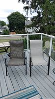 Two patio chairs shown side by side on a deck: one with black steel frame and dark gray fabric, one with aluminum frame and beige fabric.