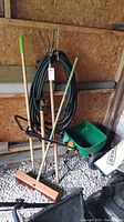 Photo showing black garden hose coiled on wall hook, broom with green handle grip, Scotts Turf Builder spreader on floor, metal weed remover and metal edger leaning against wall.