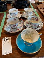 Five vintage teacups and saucers on wooden table, various floral designs and one turquoise set.