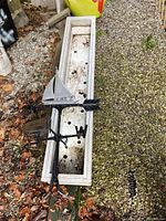 Wide-angle photo showing aluminum weather vane laying on top of the rectangular wooden planter box, both on ground with leaves and gravel around.