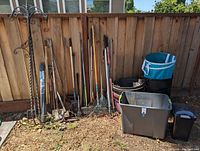 Wide shot of garden tools leaning against a wooden fence including rakes, shovels, pitchforks and a wrought iron plant hanger. On the ground are large plastic bins and pots as well as trash cans.
