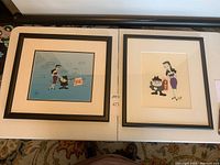 Two black framed sericels displayed side by side on a table