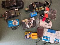 Assorted cameras and accessories on floor: film cameras, point-and-shoot cameras, film boxes, filters and straps