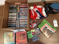 Cardboard box of approximately 70 jewel-case audio CDs, folded Union Jack and Highland Games flags, nylon CD wallet, sealed blank cassette tapes and other tapes