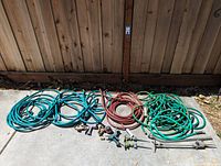 Multiple garden hoses—blue, red, and green—coiled on pavement with several hose accessories including spray nozzles, brass fittings, sprinkler heads, and metal hose stakes.