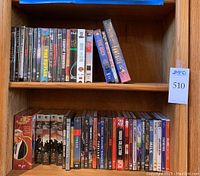 Wide view of wooden shelf with approximately 40 VHS and DVD cases including blank Fuji VHS tape and multiple movie and TV show titles.