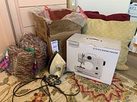 Photo showing Magicfly 12-stitch sewing machine in original box, three pillows (two red velvet, one yellow geometric), small iron, and cardboard box with yarn skeins