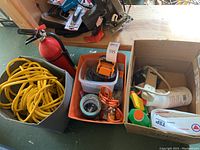 Photo showing heavy duty yellow extension cord coiled inside a box next to fire extinguisher and orange corded power stapler and other miscellaneous hardware items in boxes on wood surface.