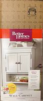 Box with Better Homes and Gardens Cottage wall cabinet image showing white cabinet with two doors, knobs, and bottom shelf.