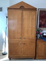 Full front view of the armoire showing caramel maple finish, triangular pediment with walnut inlay, and four door panels.