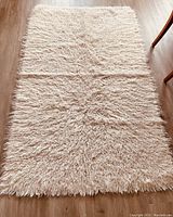 Full top view of an off-white Greek Flokati rug on wooden flooring showing long shag wool fibers and fluffy texture.