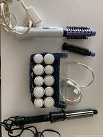 Conair hot air styling brush with two barrel brush attachments in white and lavender, Remington heated hair rollers set in blue pouch and clips, all laid flat with cords visible