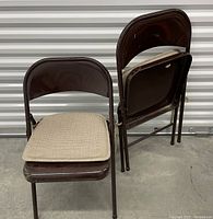 Two brown metal folding chairs with cream cushions, one chair folded, showing the shape and construction.