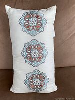 Front view of off-white throw pillow showing three embroidered floral-like circular patterns in teal and reddish-brown thread on a rectangular fabric.
