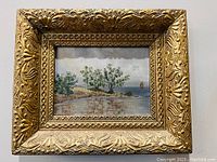 Full view of framed oil painting showing coastal landscape scene with trees and a sailboat, within an ornate gold frame.