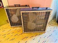 Two vintage box fans placed side by side on a patterned carpet near a pink wall. They have beige metal frames and black top panels with handles.