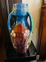 Front view of tall pottery vase showing blue glaze top and handles with reddish brown paneled body and raised botanical designs.