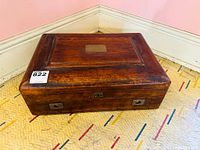 Closed antique wooden flatware chest showing top, front edge and side handle, with a smooth dark brown finish and central brass plate on lid.