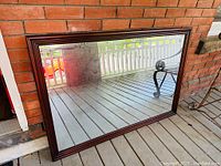 Photo showing entire large mirror leaning against a brick wall on a porch with clear glass reflecting surroundings.