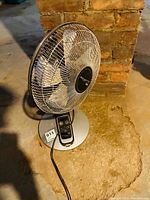 Front view of the oscillating table fan showing fan blades, grille, control panel, and power cord on concrete floor against brick wall.