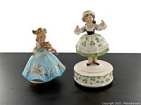 Front view of both figurine music boxes together on black surface showing Irish-themed girl in white dress with shamrocks and girl in blue floral dress playing violin.