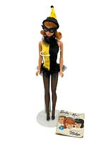 Full front view of the 1964 Midge doll wearing black and yellow Masquerade outfit with mask and hat, with original Midge book placed at feet.