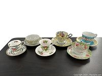 Six vintage fine china cups and saucers displayed on black surface showing varied floral patterns and colors.