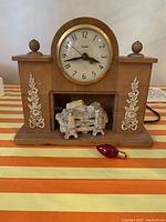 Front view of wooden mantel clock with carved decorations, round clock face and faux fireplace with loose log set and red bulb placed in front.