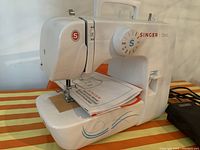 Front and side view of the white Singer Start sewing machine with user manual on the base and foot pedal beside it.