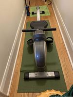 Rowing machine positioned on a green yoga mat within a narrow hallway, showing the handlebar and seat from the front view.