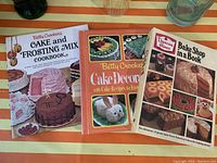 Photo showing three vintage dessert cookbooks: Betty Crocker's Cake and Frosting Mix Cookbook, Betty Crocker's Cake Decorating Cookbook, and Duncan Hines Bake Shop in a Book, arranged on a striped surface with glasses and bottles nearby.