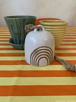 Photo showing two ceramic planters and a ceramic bell with rope on a striped surface.