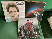 Three vintage LP records laid out together. Huey Lewis and the News 'Picture This' front cover showing male portrait, Van Morrison 'Beautiful Vision' cover with Earth and light trails, Donnie Iris 'The High and the Mighty' cover with artist in red jacket against city background.
