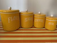 Front view of four yellow kitchen canisters labeled Flour, Sugar, Coffee, Tea on striped surface.