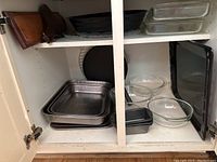 Photo of cabinet shelf with assorted baking pans including metal roasting pans, glass casserole dishes, glass mixing bowls, and black metal baking pans.