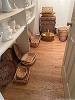 Photo showing a large rectangular wicker hamper with lid, several hand-held and flat woven trays and baskets in various sizes and shapes on a wooden floor.